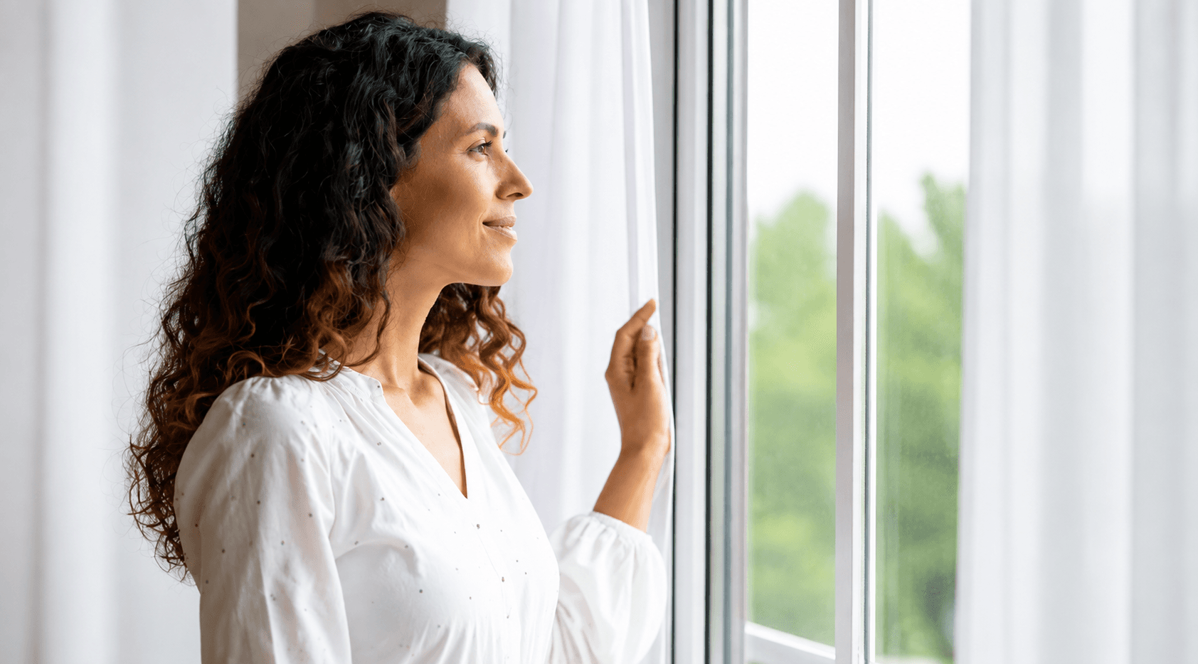 A woman smiling as she looks out of a bright window, holding a sheer white curtain